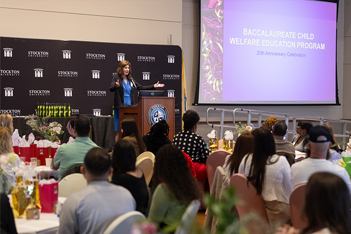 Diane Falk stands behind a podium while she addresses a crowd seated at round tables in the Campus Center Event Room