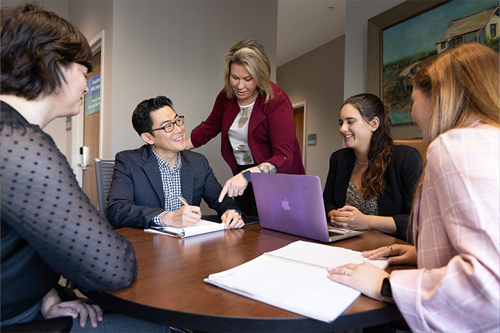 A faculty member stands and points over a seated student's shoulder, with other students seated around the table
