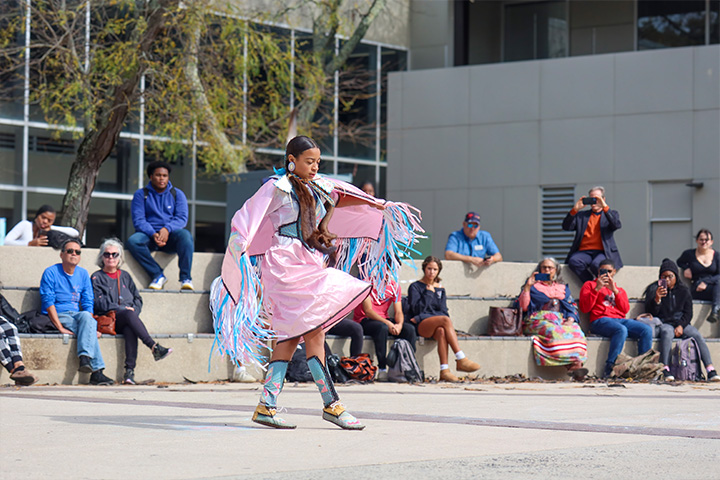 A woman wearing a traditional Indigenous dress of light pink with blue fringe dances outside Stockton's main academic building