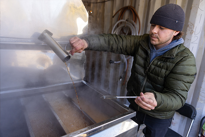 Ryan Hagerty wears a black winter hat while pouring fresh, hot maple syrup into a vat.
