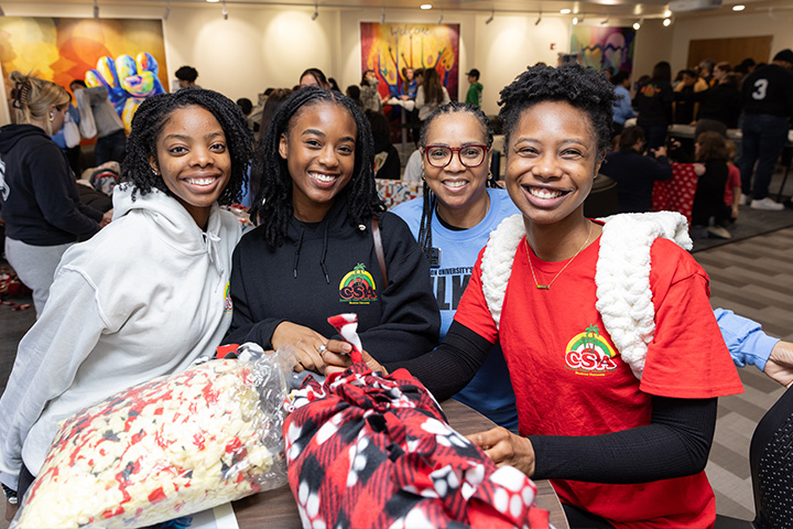 Four women smile while standing around a table top filled with fabric scraps used to make blankets