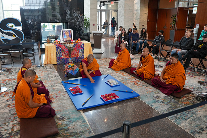 Six Tibetan monks in orange wraps sit on the floor in the Campus Center Grand Hall with a large blue square board that will serve as the base for the mandala