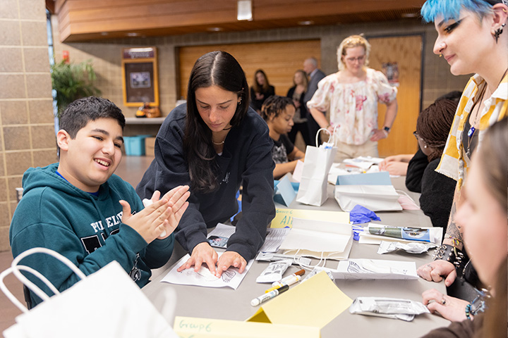 A young student in a Philadelphia Eagles sweatshirt molds modeling clay while college students assist