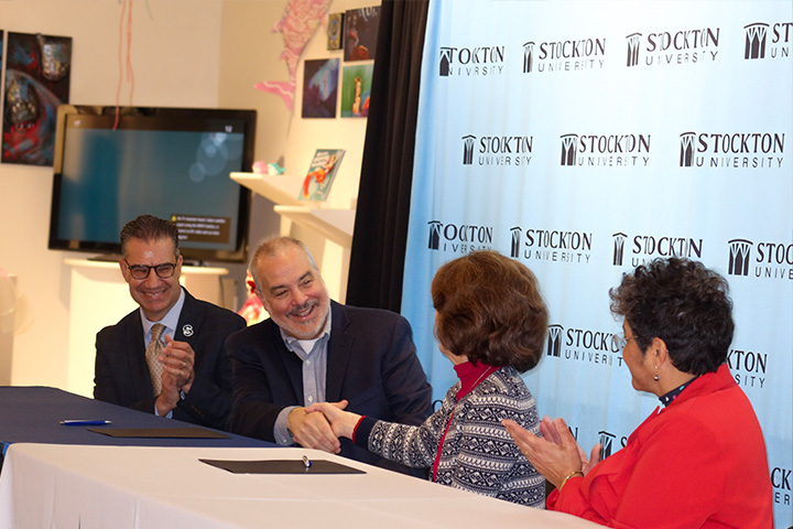Four people sitting at a long table, with the two individuals in the middle shaking hands