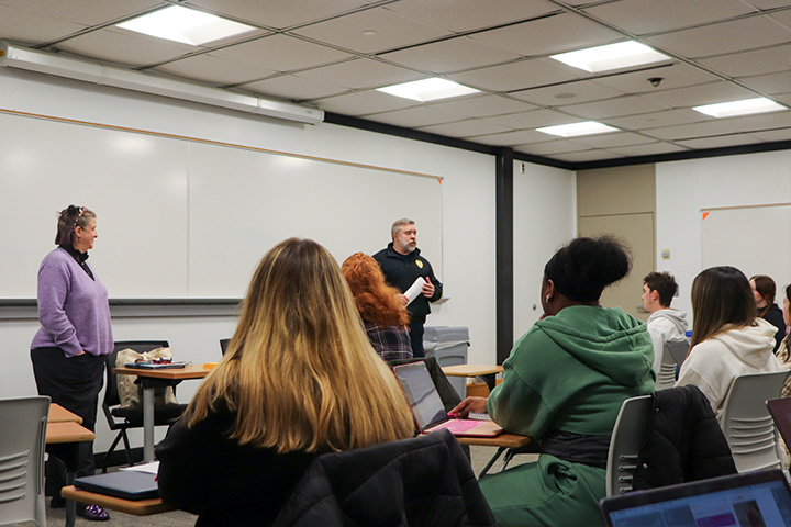 An adult stands in the front of a classroom talking to students sitting in desks
