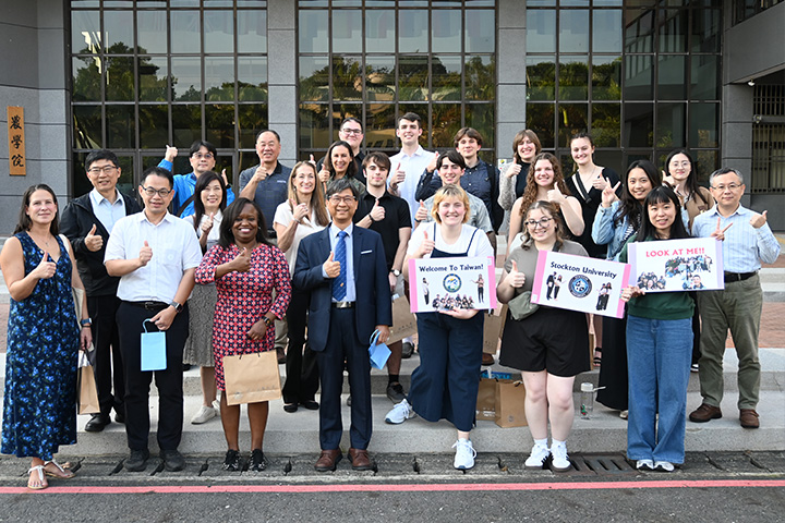 A group of Stockton faculty and students stand outside National Chiayi University in Taiwan holding their thumbs up in a positive gesture