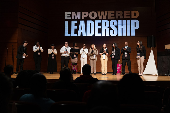 A group of students stand on stage in the Campus Center Theatre with a logo reading Empowered Leadership screened behind them