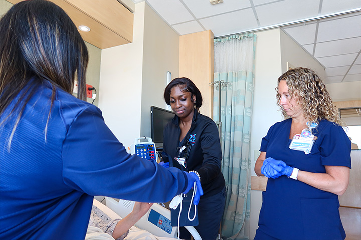 Amiesa Paasewe in dark blue scrubs works with two other nurses to care for a patient in a hospital bed