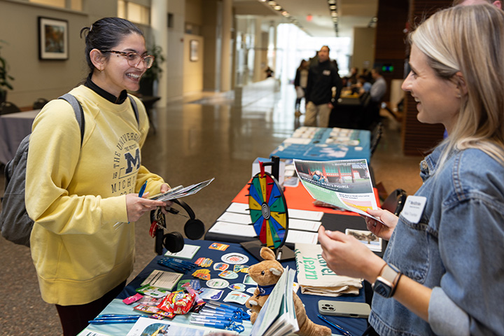 A student in a yellow sweatshirt stands on one side of a table chatting with another student in a denim shirt on the other side of the table