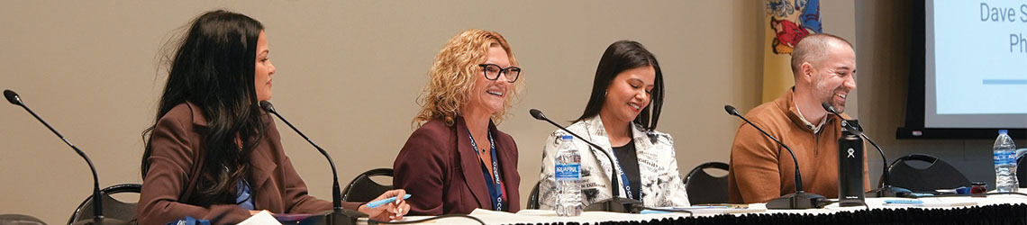 Four panelists sit at a long table with tall microphones