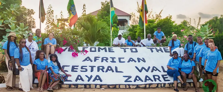 A group of students gathered outdoors in front of a large banner that reads “WELCOME TO THE AFRICAN ANCESTRAL WALL LAYA ZAARE”. 