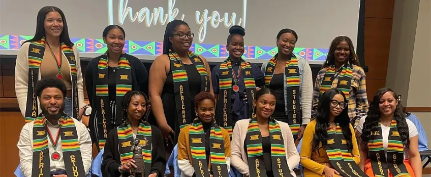 A group of Stockton students at a graduation ceremony, each wearing a colorful stole with stripes of green, yellow, red, and black.
