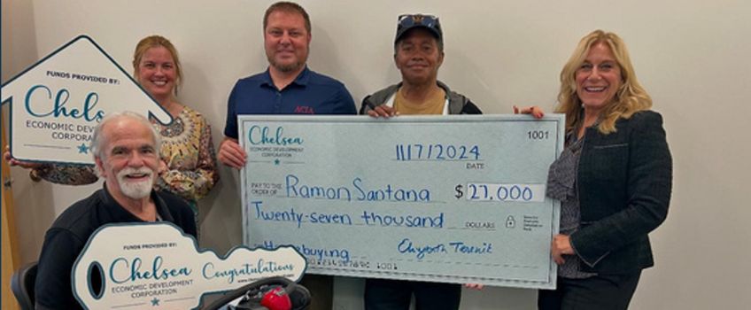 Elizabeth Terenik and Chelsea EDC staff present resident, Ramon Santana, with an oversized check. Mr. Santana sits at the front holding a giant key.  