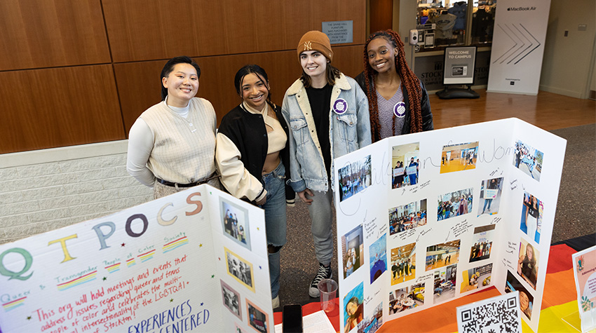 E-board members of QTPOCS and Womens Co, standing behind their table and signs during the celebration.