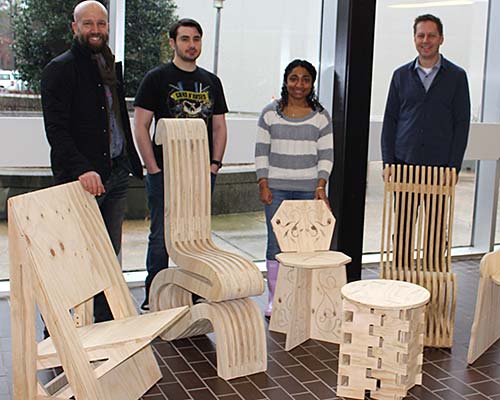 male professor, two male students and female student stand behind wooden chairs they created