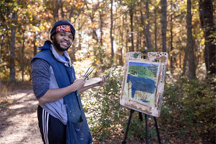 A student wearing a snow hat stands in front of an easel with a painting of a lake while standing outside along a wooded path