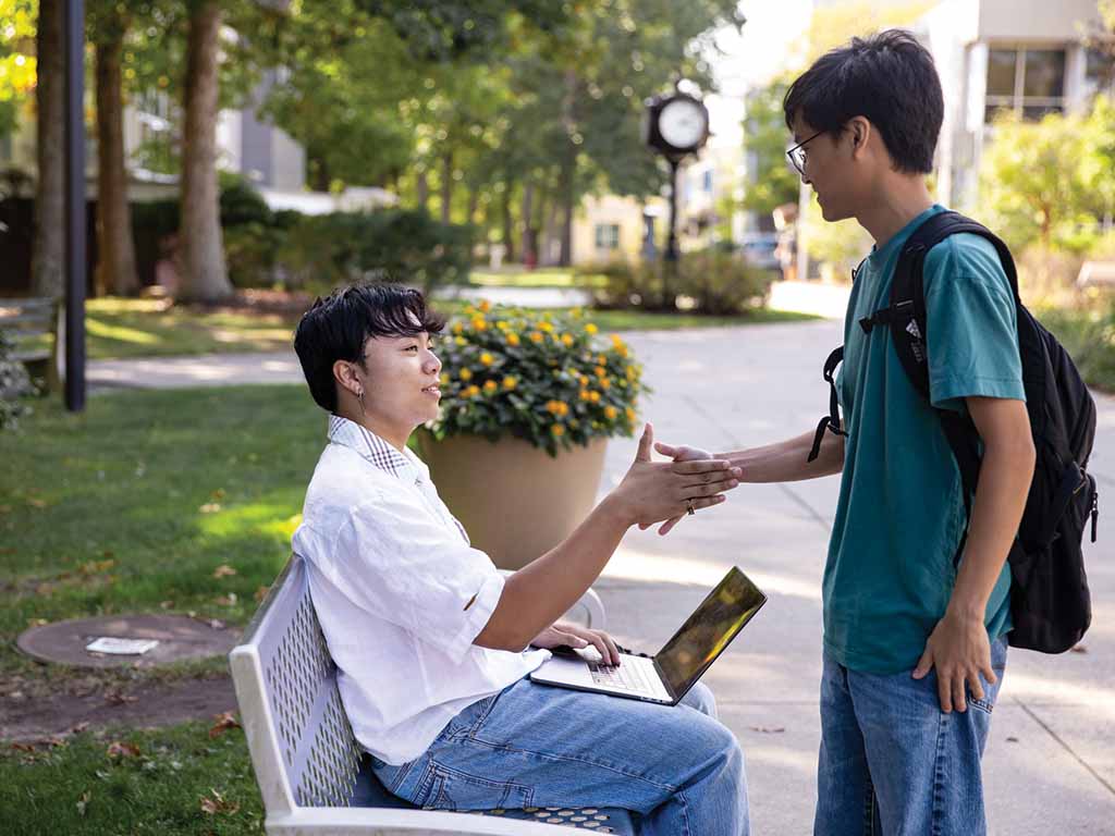 A Stockton University student sitting on a bench with a laptop shakes hands with another Stockton student standing with a backpack