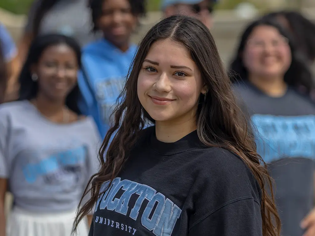 Stockton University student smiling outdoors in a Stockton sweatshirt, with classmates in the background.