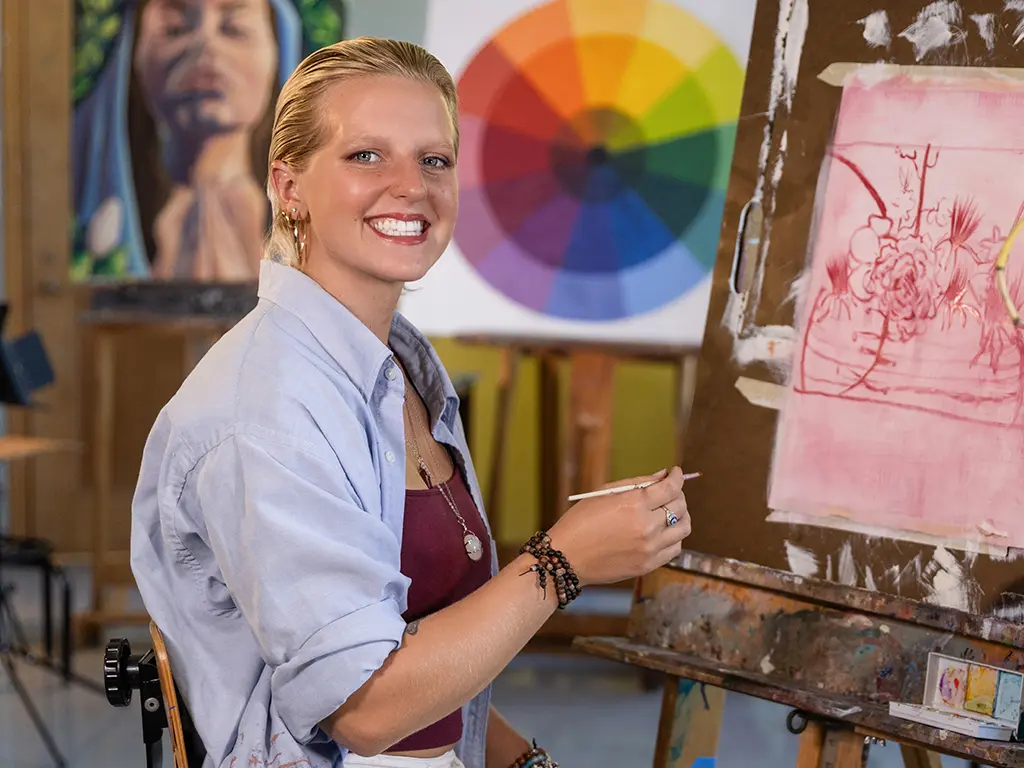 Stockton University art student smiling while painting at an easel in a studio, with a color wheel and portrait artwork in the background.