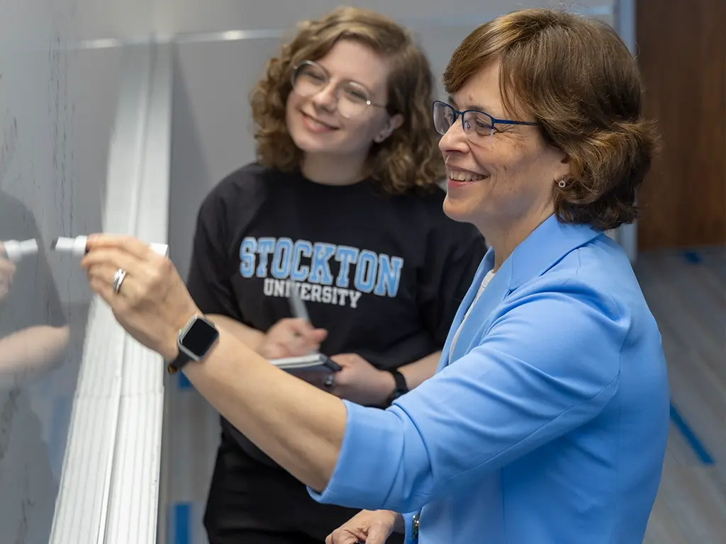 Stockton University professor writing on a whiteboard while working with a student in class.