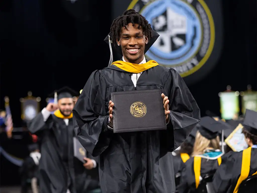 Stockton University graduate in cap and gown smiling while holding a diploma cover during commencement.