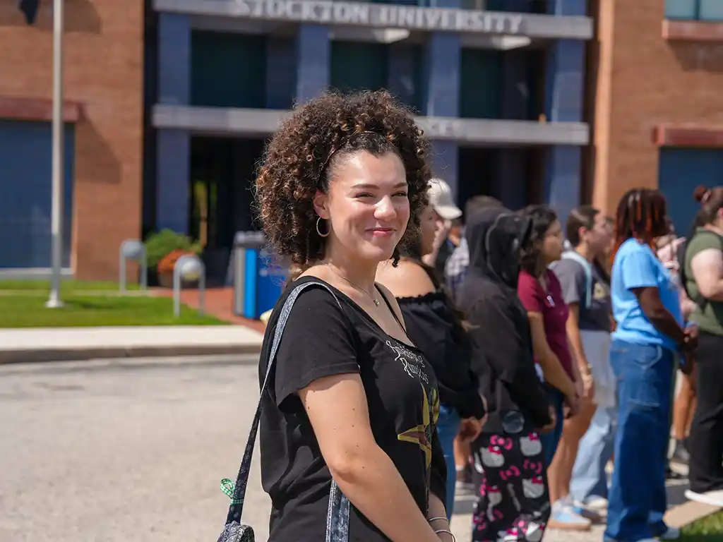 Student smiling outside Stockton University during the Hispanic Heritage Month Flag Raising, with a group of peers gathered in the background.