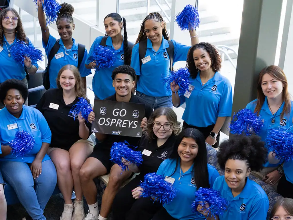 Stockton University student leaders in matching shirts holding blue pom-poms and a “Go Ospreys” sign.