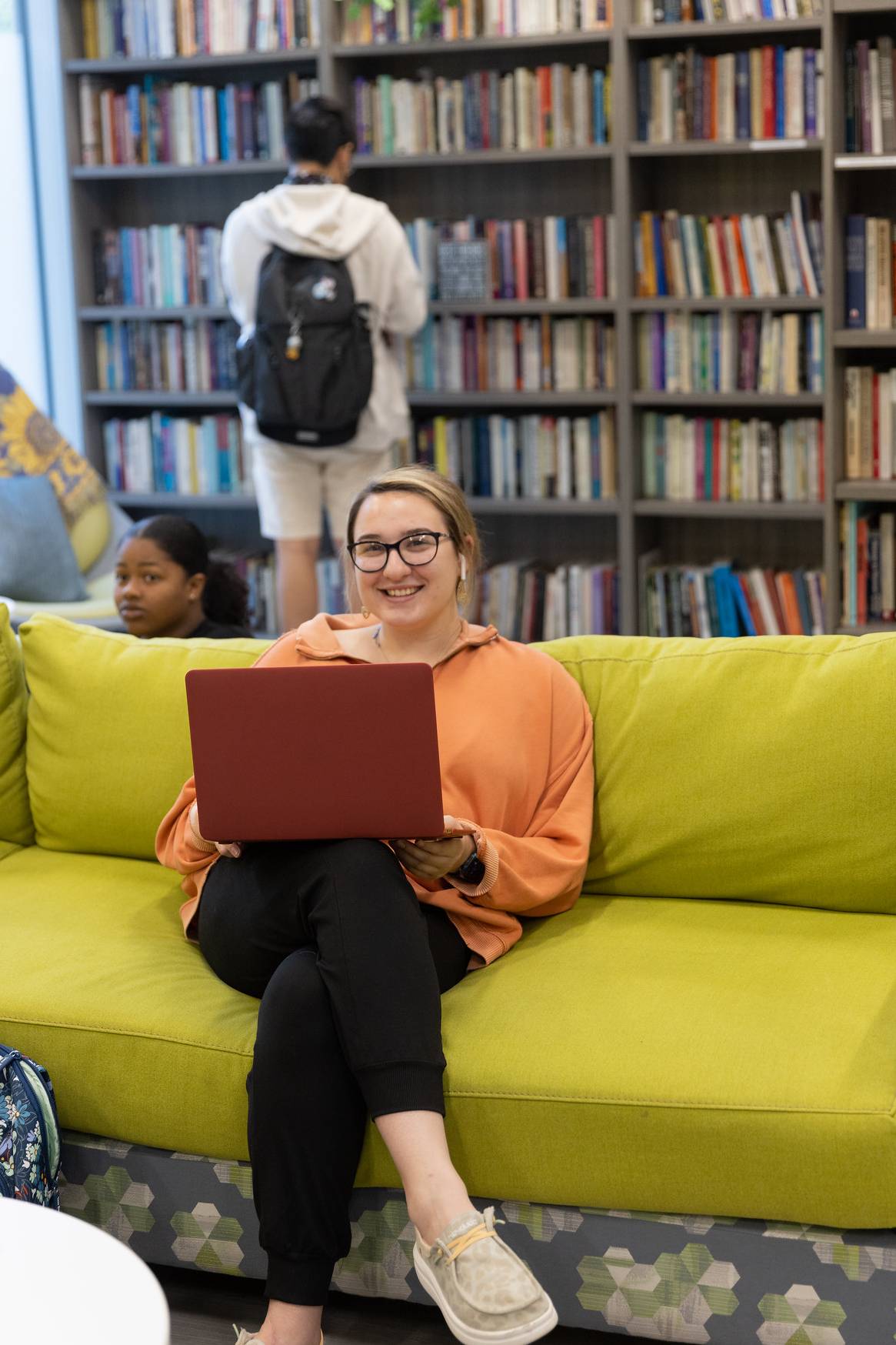 Girl using laptop on green couch with student looking at library.